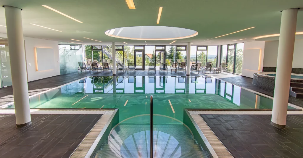 Espace piscine bien-être de l'hôtel de bien-être Hohenrodt, vue d'ensemble à la lumière du jour avec chaises longues et grande façade vitrée